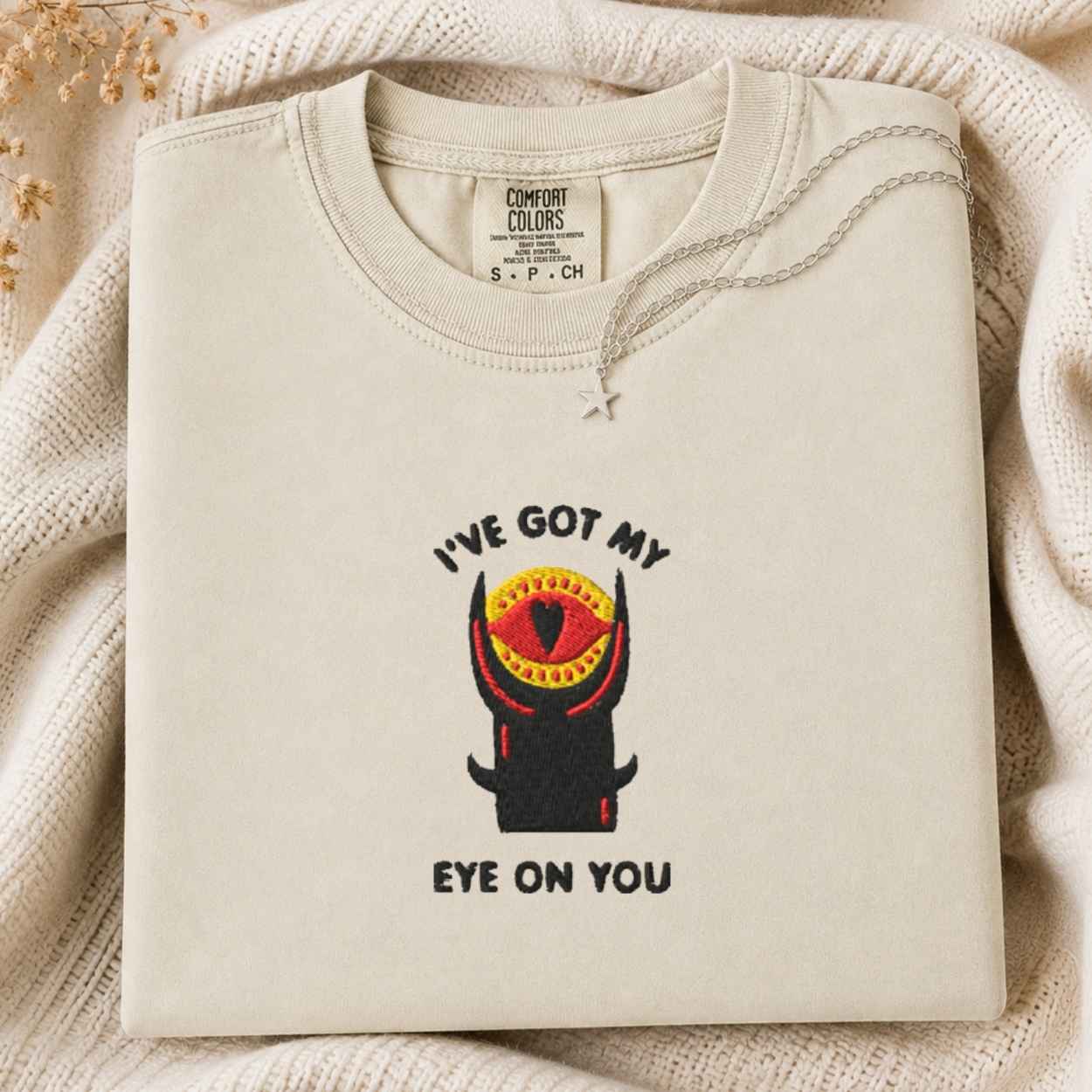 A folded cream tshirt with a graphic of a horned tower and the text 'I'VE GOT MY EYE ON YOU' is laid on a textured cream knit blanket. The neck label reads 'COMFORT COLORS' and 'S.
