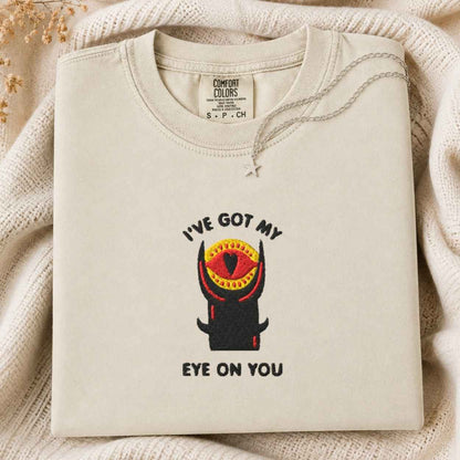 A folded cream tshirt with a graphic of a horned tower and the text 'I'VE GOT MY EYE ON YOU' is laid on a textured cream knit blanket. The neck label reads 'COMFORT COLORS' and 'S.