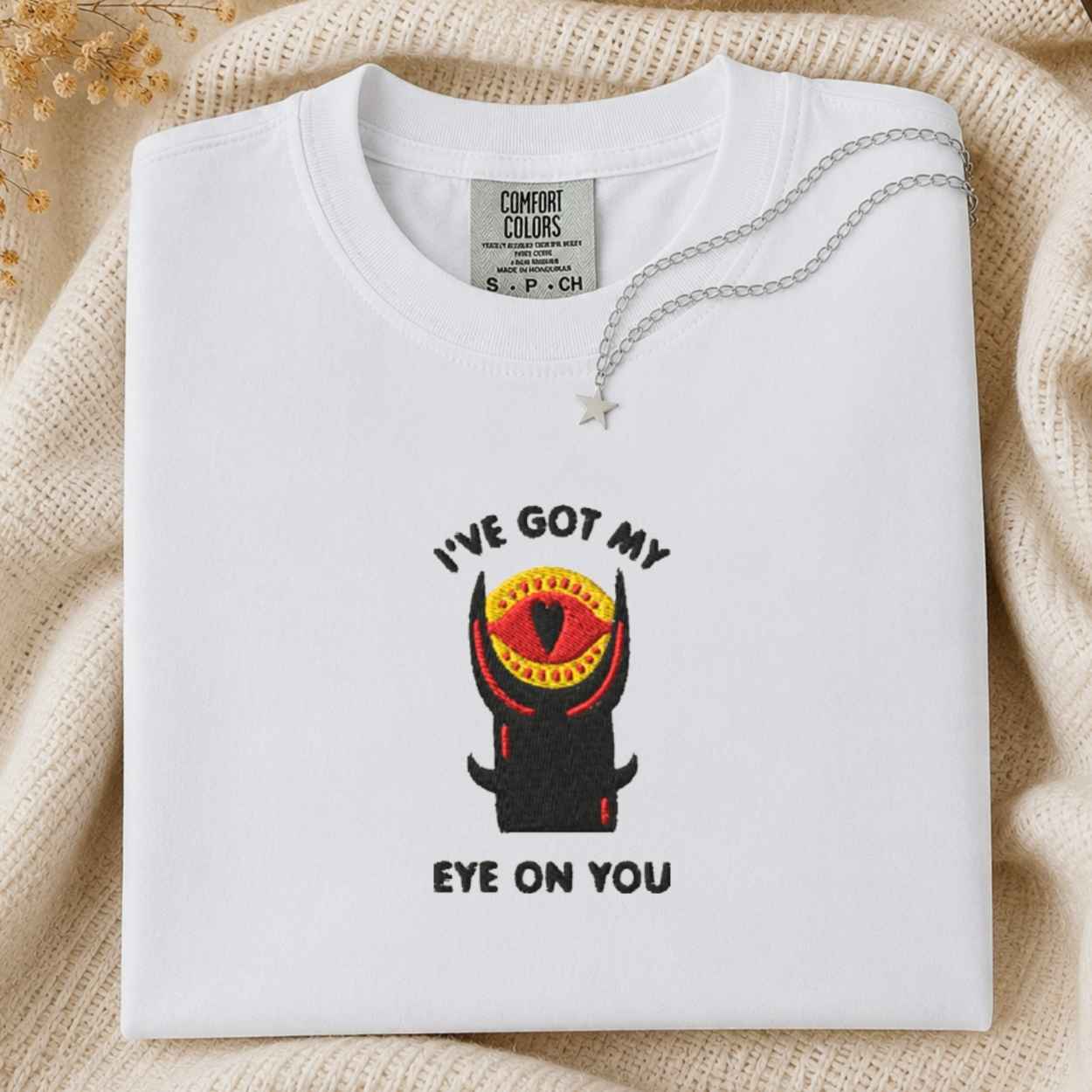 A folded white t-shirt with a graphic of a black horned tower with a red and yellow eye, accompanied by the text 'I'VE GOT MY EYE ON YOU'. A silver necklace is draped over the collar.