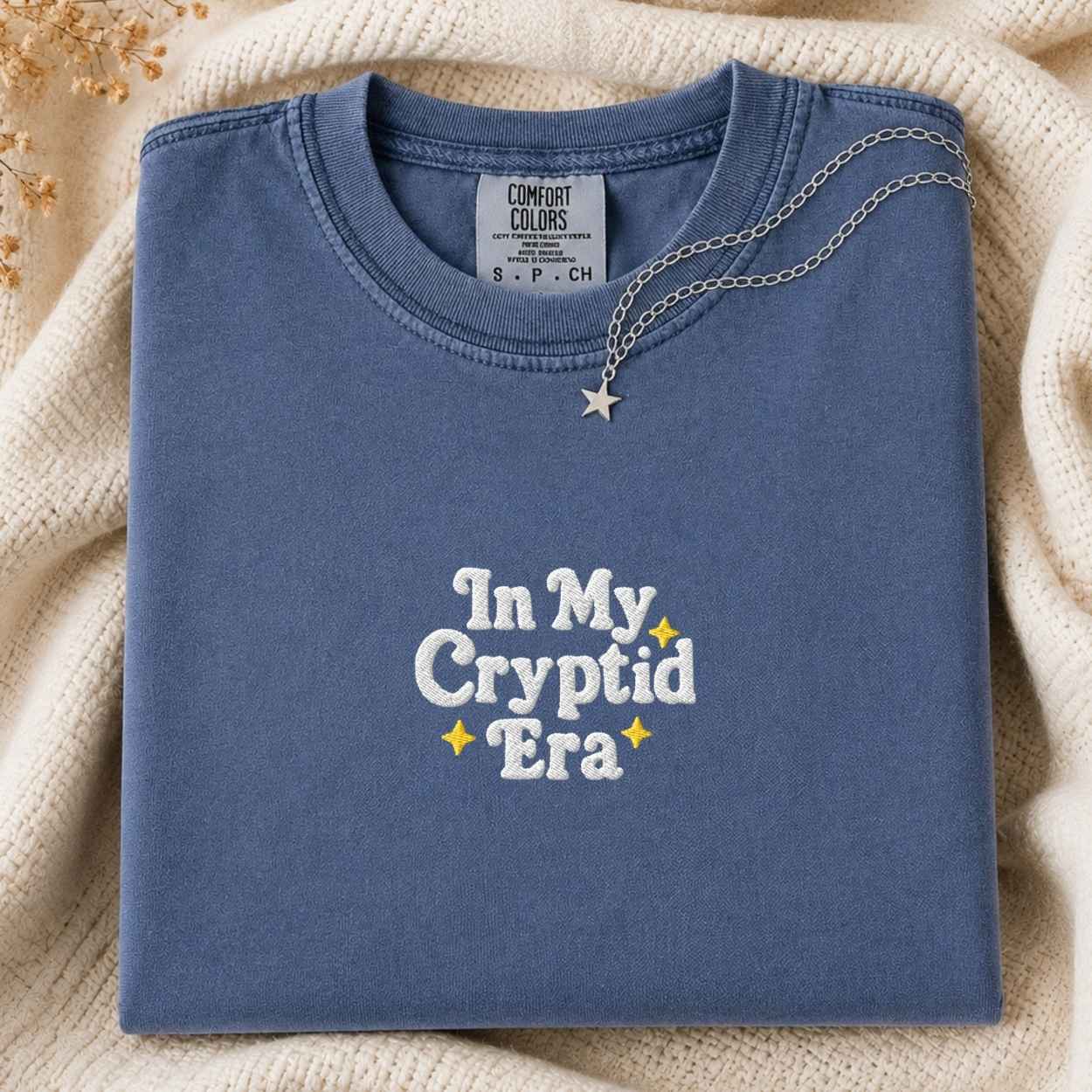 A folded slate blue t-shirt with 'In My Cryptid Era' embroidered in white and yellow on the front, styled on a cozy cream knit blanket with a silver star necklace draped over the collar.