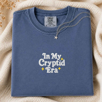 A folded slate blue t-shirt with 'In My Cryptid Era' embroidered in white and yellow on the front, styled on a cozy cream knit blanket with a silver star necklace draped over the collar.
