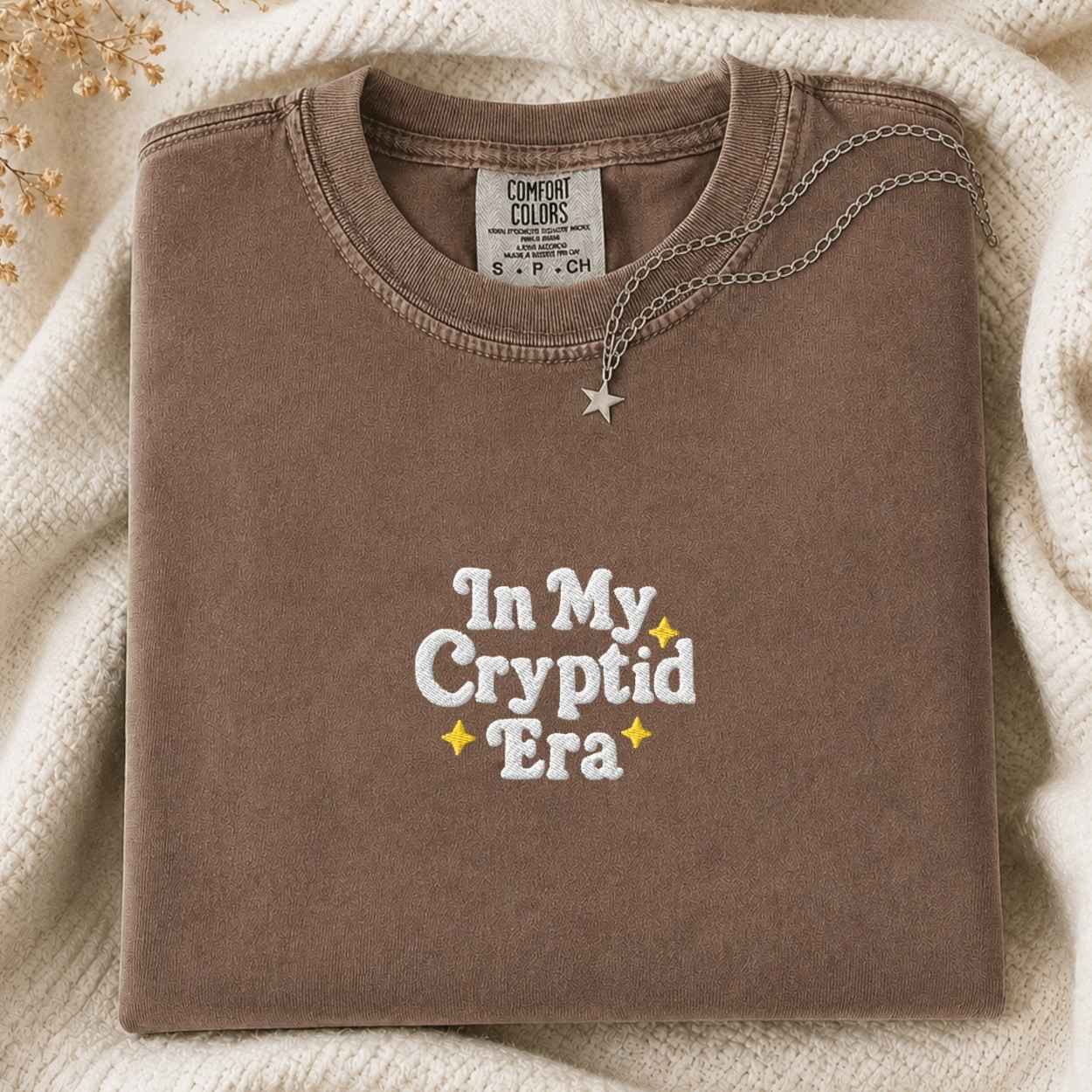 A folded brown t-shirt with 'In My Cryptid Era' embroidered on the front, styled on a cream knit blanket with a silver necklace.