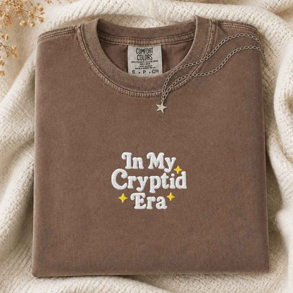 A folded brown t-shirt with 'In My Cryptid Era' embroidered on the front, styled on a cream knit blanket with a silver necklace.