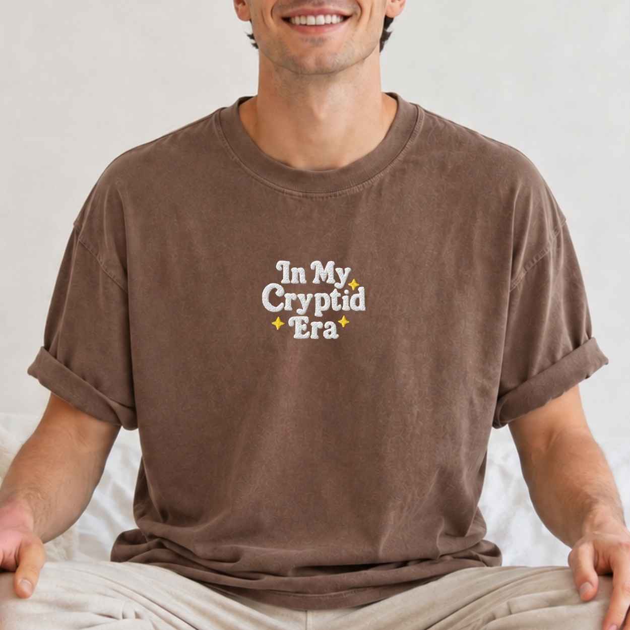 A man wearing a brown crewneck t-shirt with rolled-up sleeves and beige pants, sitting cross-legged. The shirt features the text 'In My Cryptid Era' printed on the chest in white with small yellow star accents.