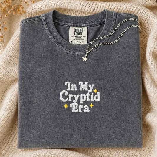 A folded charcoal gray Comfort Colors t-shirt with 'In My Cryptid Era' embroidered in white and yellow on the front, styled on a beige knit blanket with a silver star necklace draped over the collar.