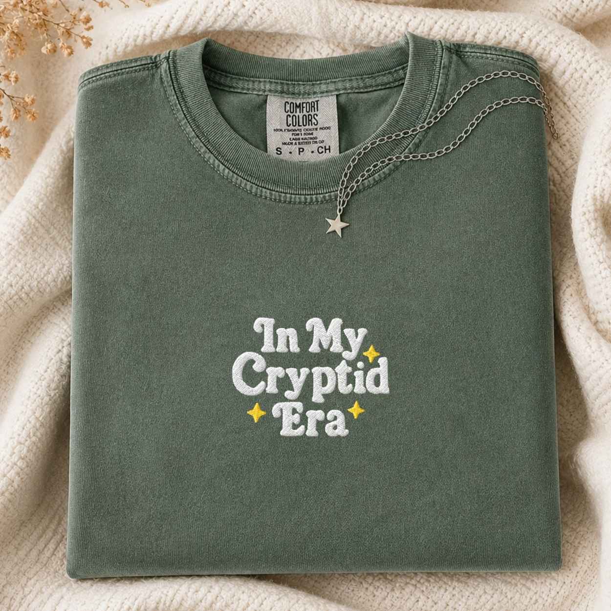 A folded sage green Comfort Colors t-shirt with 'In My Cryptid Era' embroidered on the front, styled on a cream knit blanket with a silver star necklace.