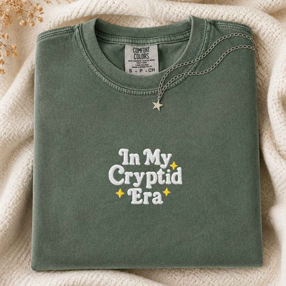 A folded sage green Comfort Colors t-shirt with 'In My Cryptid Era' embroidered on the front, styled on a cream knit blanket with a silver star necklace.