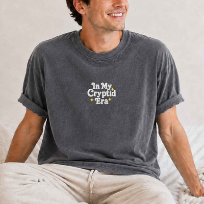 A man wearing a dark gray washed t-shirt with the text 'In My Cryptid Era' printed on the chest, sitting on a bed.