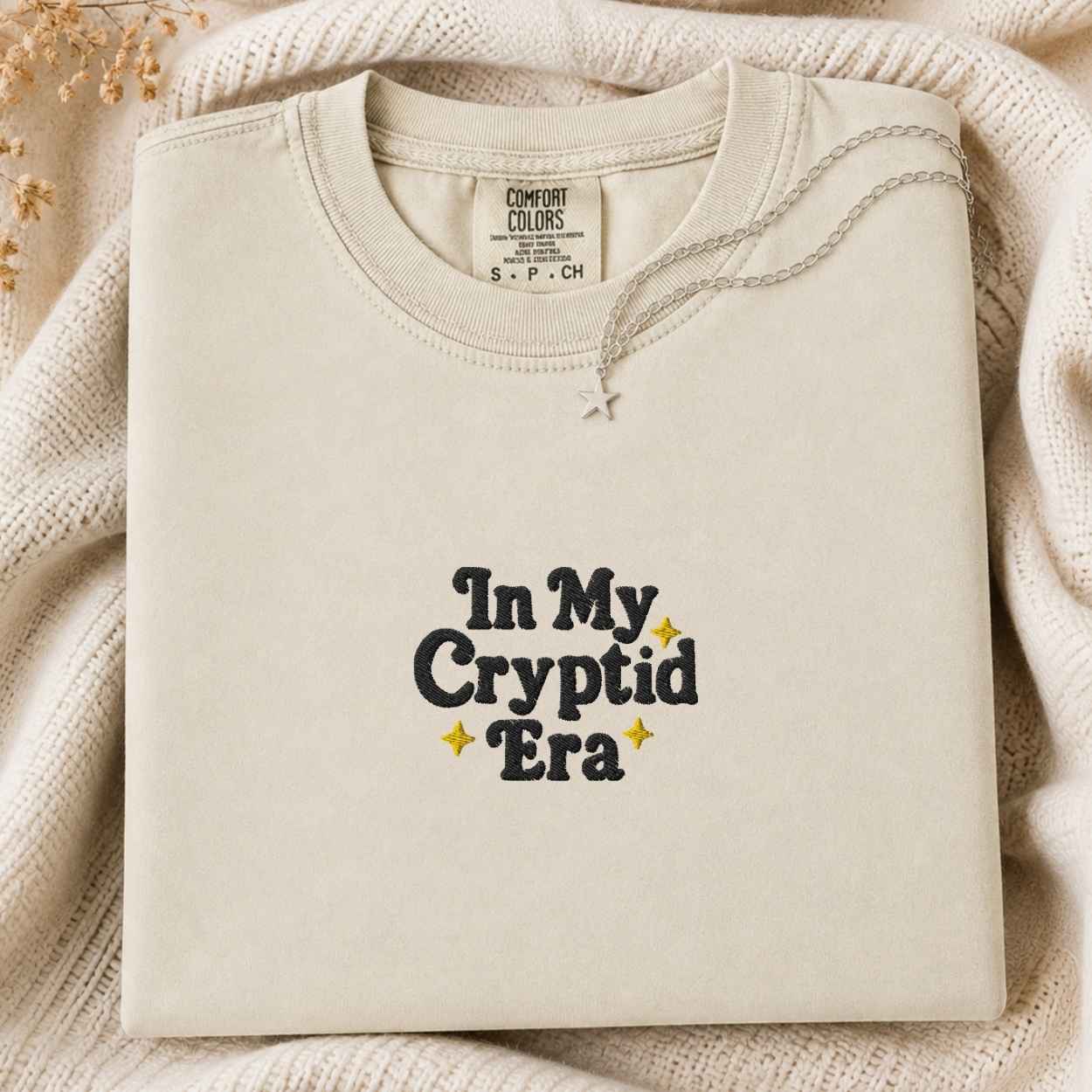 A folded cream-colored crewneck sweatshirt with the text 'In My Cryptid Era' printed on the front in black and yellow, styled on a cozy cream knit blanket with dried flowers.