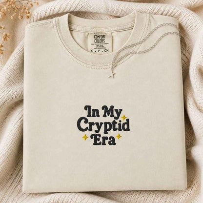 A folded cream-colored crewneck sweatshirt with the text 'In My Cryptid Era' printed on the front in black and yellow, styled on a cozy cream knit blanket with dried flowers.