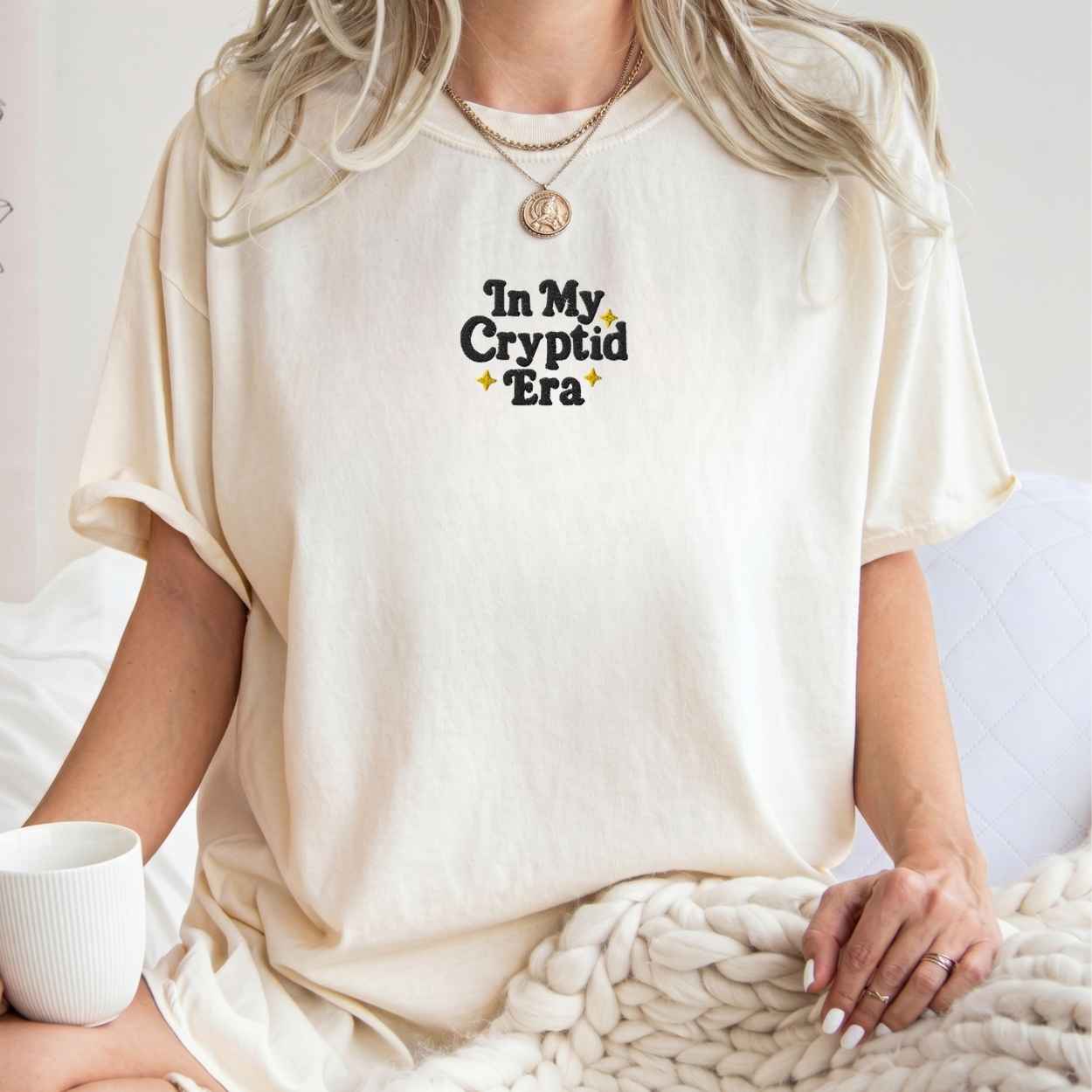A woman wearing an oversized cream-colored t-shirt with the text 'In My Cryptid Era' printed on the chest in black and yellow. She is sitting on a bed with a chunky knit blanket and a white mug nearby.