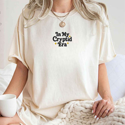 A woman wearing an oversized cream-colored t-shirt with the text 'In My Cryptid Era' printed on the chest in black and yellow. She is sitting on a bed with a chunky knit blanket and a white mug nearby.