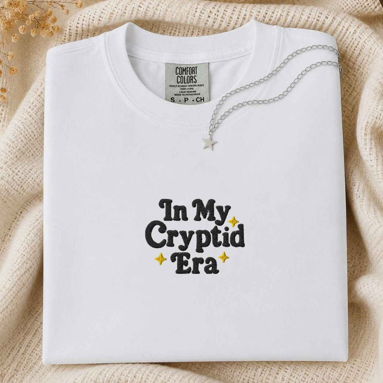A folded white Comfort Colors t-shirt with 'In My Cryptid Era' printed in black and yellow on the front, styled on a beige knit blanket with a silver necklace.