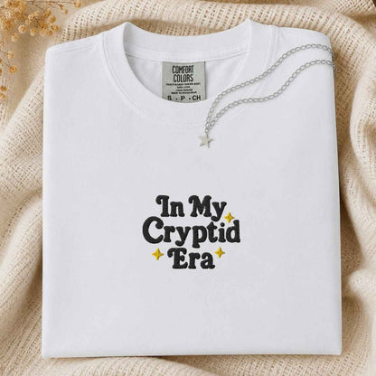 A folded white Comfort Colors t-shirt with 'In My Cryptid Era' printed in black and yellow on the front, styled on a beige knit blanket with a silver necklace.