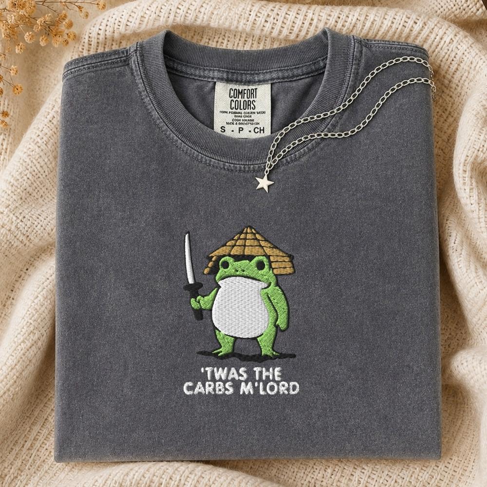 A folded dark gray t-shirt with a green frog character embroidered on the front, text is it was the carbs my lord, styled on a beige knit blanket with a silver necklace draped over the collar.