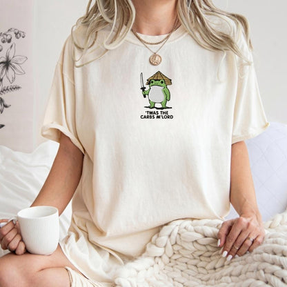 A woman wearing a cream-colored oversized t-shirt with a green cartoon frog graphic holding a sword and the text 'Twas the carbs my lord'. She is sitting in a bedroom setting with a white mug and a chunky knit blanket.