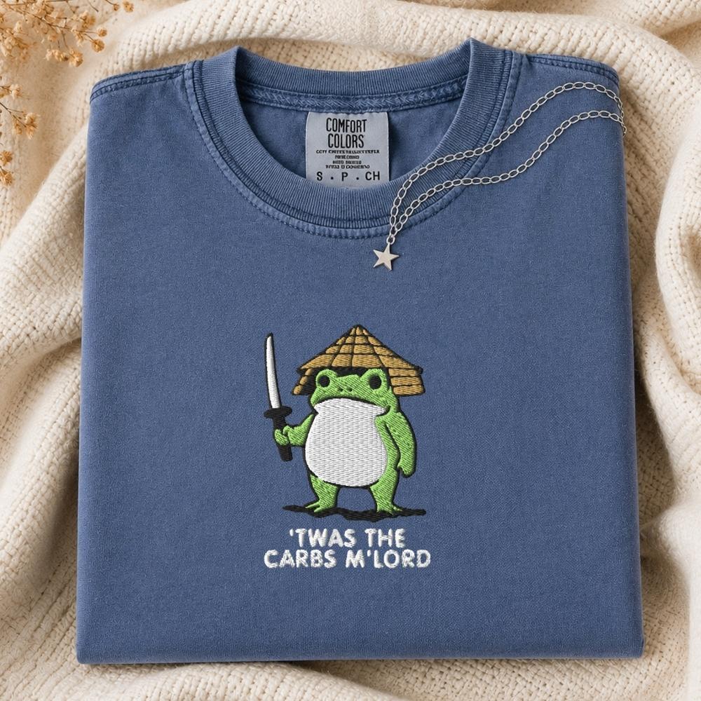 A folded slate blue t-shirt with a green cartoon frog holding a sword and wearing a straw hat, with the text 'TWAS THE CARBS M'LORD' printed below. The shirt is styled on a cream-colored knit blanket with a silver necklace draped over the collar.
