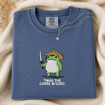 A folded slate blue t-shirt with a green cartoon frog holding a sword and wearing a straw hat, with the text 'TWAS THE CARBS M'LORD' printed below. The shirt is styled on a cream-colored knit blanket with a silver necklace draped over the collar.