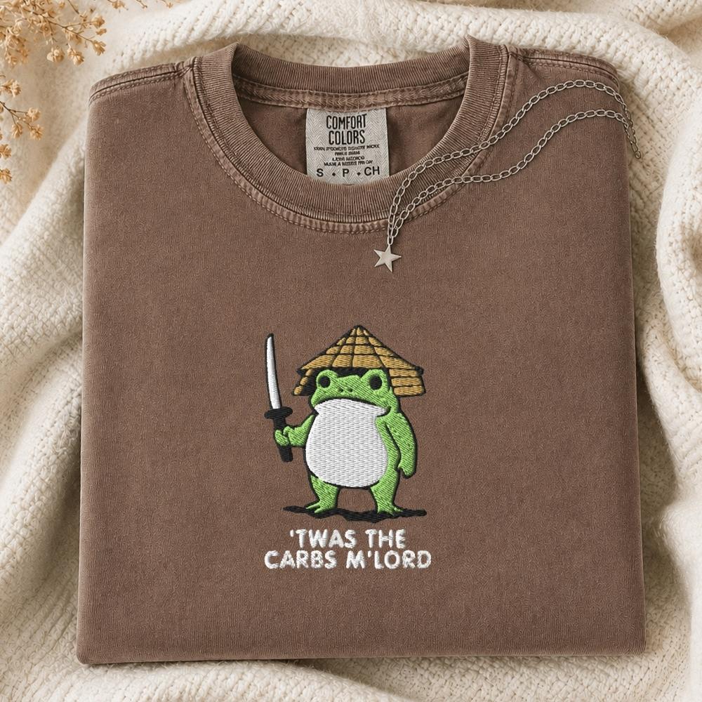 A folded brown Comfort Colors t-shirt featuring a green cartoon frog dressed as a samurai with the text 'TWAS THE CARBS M'LORD' printed on the front. The shirt is styled on a cream knit blanket with a silver necklace draped over the collar.
