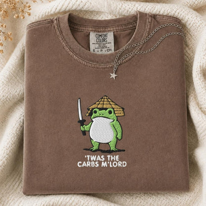 A folded brown Comfort Colors t-shirt featuring a green cartoon frog dressed as a samurai with the text 'TWAS THE CARBS M'LORD' printed on the front. The shirt is styled on a cream knit blanket with a silver necklace draped over the collar.