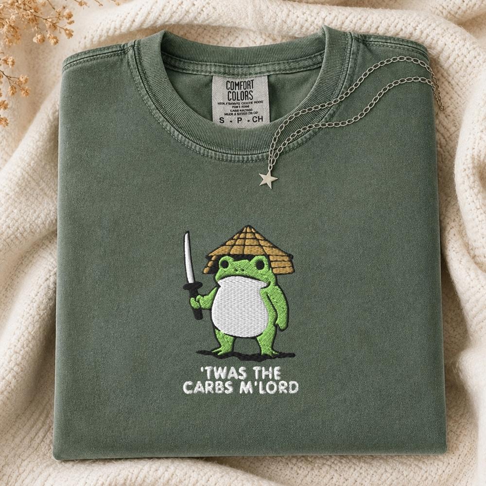 A folded sage green Comfort Colors t-shirt featuring a cartoon frog character wearing a straw hat and holding a sword, with the text 'TWAS THE CARBS M'LORD' printed below. The shirt is styled on a cream-colored knit blanket with a silver necklace draped over the collar.