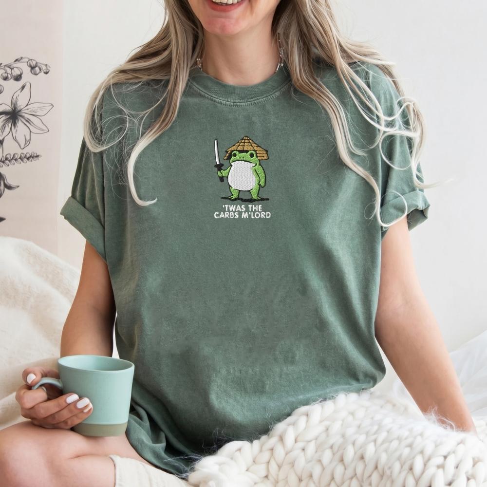 A woman wearing a sage green graphic t-shirt with a cartoon green frog holding a sword and the text 'T WAS THE CARBS M' LORD. She is holding a mint green mug and sitting on a bed with a chunky knit blanket.