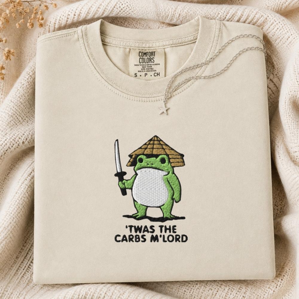A folded cream-colored tshirt with a green cartoon frog holding a sword and wearing a straw hat, with the text 'TWAS THE CARBS M'LORD' printed below. The sweatshirt is displayed on a cozy beige knit blanket.