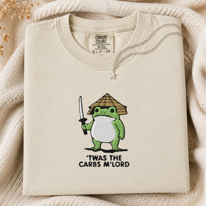 A folded cream-colored tshirt with a green cartoon frog holding a sword and wearing a straw hat, with the text 'TWAS THE CARBS M'LORD' printed below. The sweatshirt is displayed on a cozy beige knit blanket.