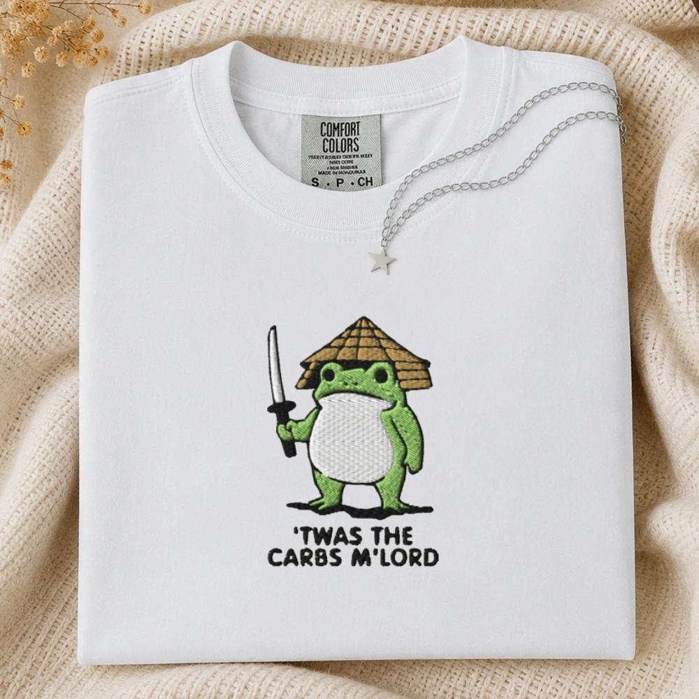 A folded white Comfort Colors t-shirt with a green cartoon frog holding a sword and a straw hat, with the text 'TWAS THE CARBS M'LORD' printed below. The shirt is styled on a beige knit blanket with a silver necklace draped over the collar.