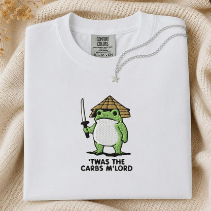 A folded white Comfort Colors t-shirt with a green cartoon frog holding a sword and a straw hat, with the text 'TWAS THE CARBS M'LORD' printed below. The shirt is styled on a beige knit blanket with a silver necklace draped over the collar.