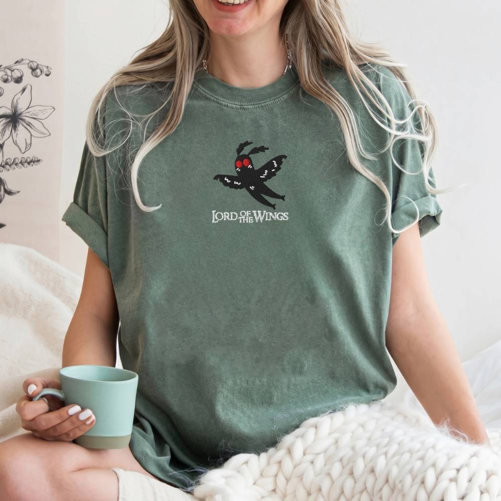 A woman wearing a sage green graphic t-shirt with a black spider character and the text 'Lord of the Wings' on the chest, holding a mint green mug while sitting on a bed with a chunky knit blanket.