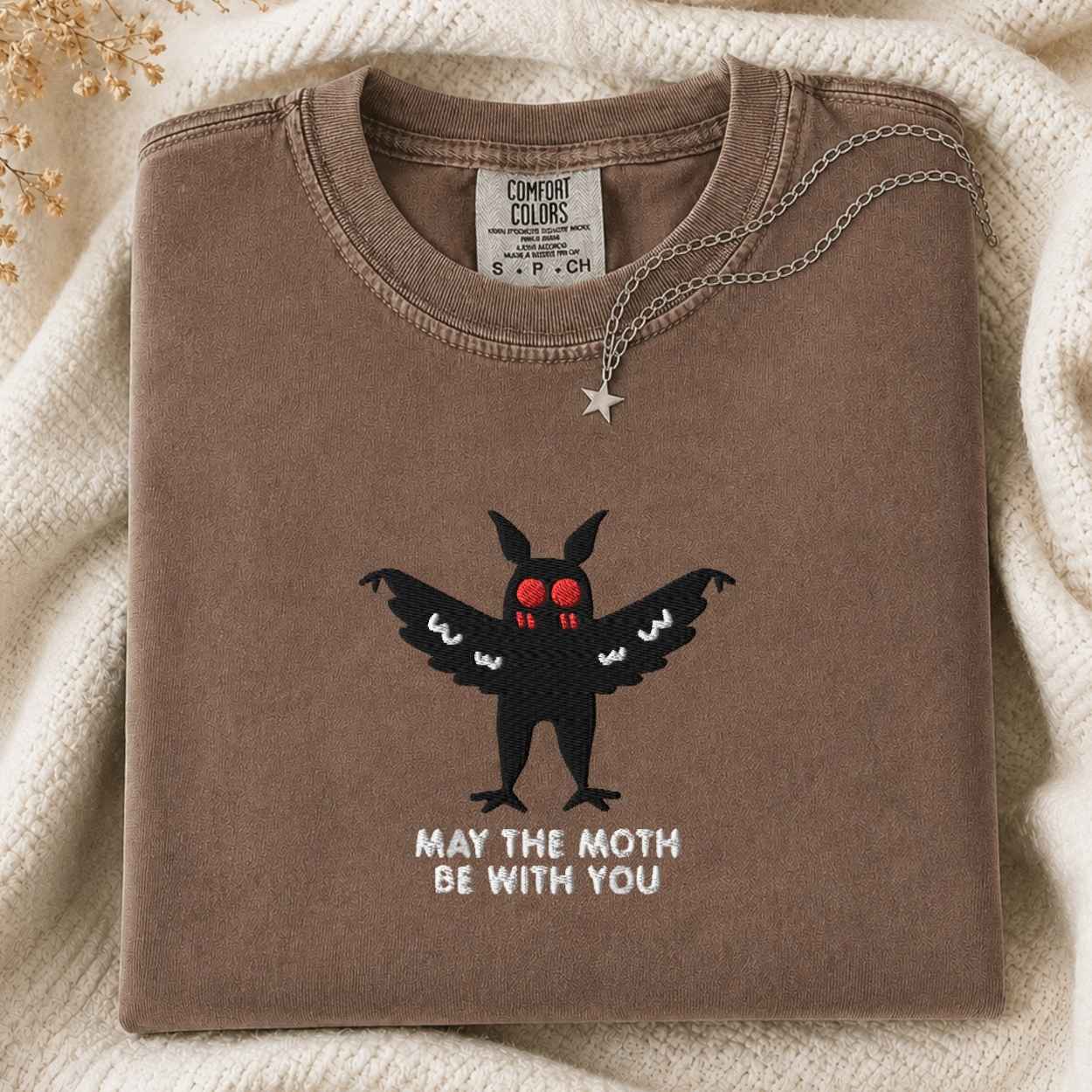 A folded taupe-brown t-shirt with a black moth graphic and the text 'MAY THE MOTH BE WITH YOU' on the front, styled on a cream knit blanket with a silver necklace.