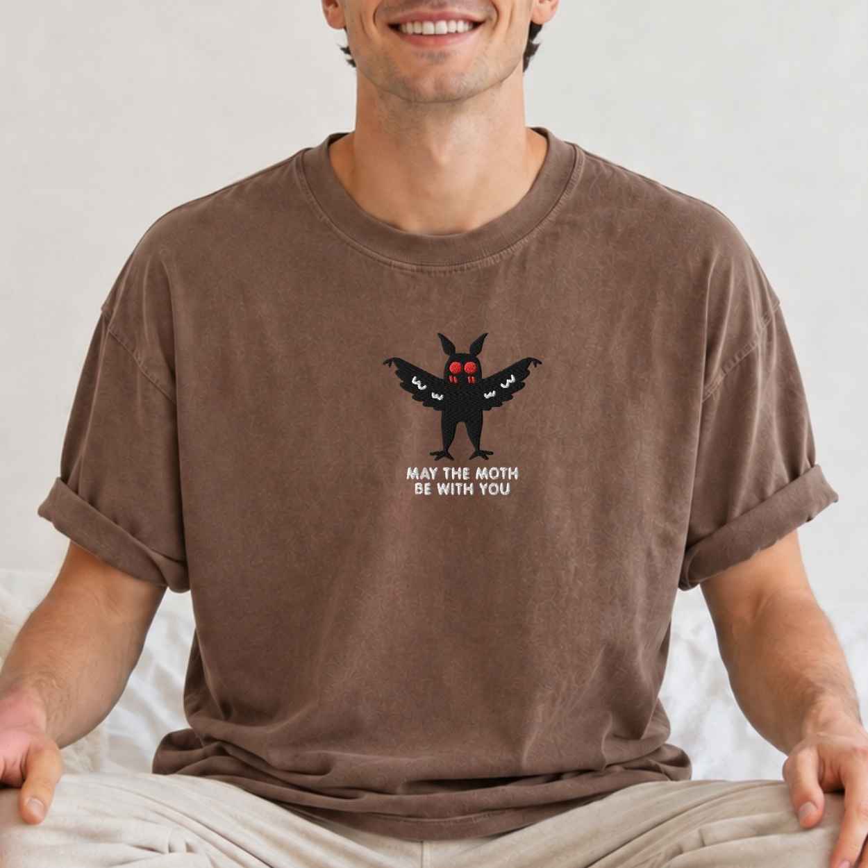 A man wearing a brown crewneck t-shirt with a graphic of a black moth with red eyes and the text 'MAY THE MOTH BE WITH YOU' on the chest, sitting against a light background.