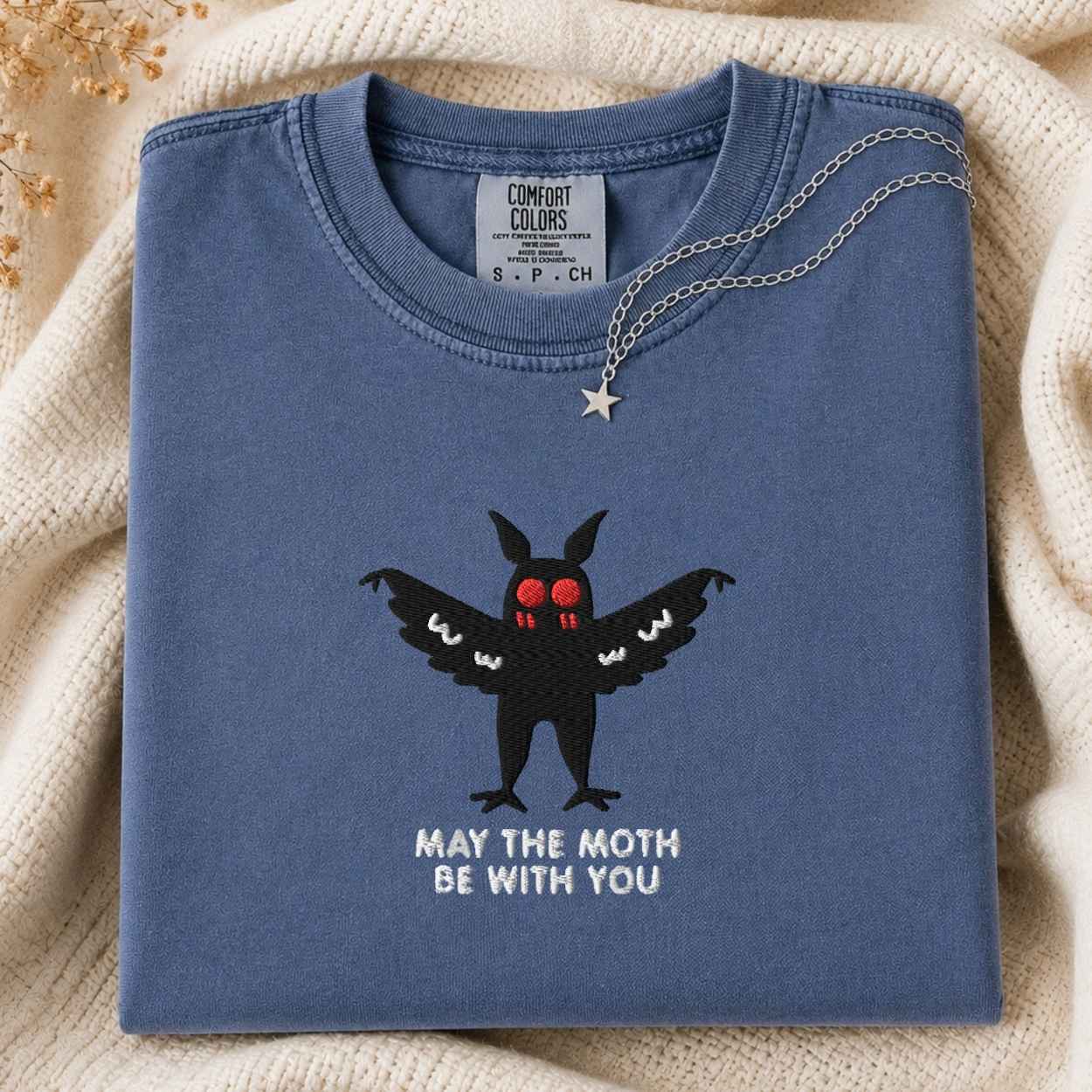 A folded slate blue t-shirt with a black mothman graphic and the text 'MAY THE MOTH BE WITH YOU' on the front, styled on a beige knit blanket with a silver necklace draped over the collar.