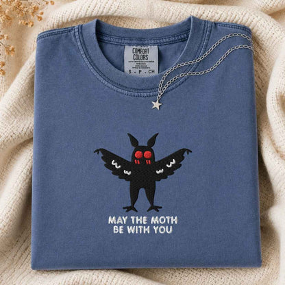 A folded slate blue t-shirt with a black mothman graphic and the text 'MAY THE MOTH BE WITH YOU' on the front, styled on a beige knit blanket with a silver necklace draped over the collar.