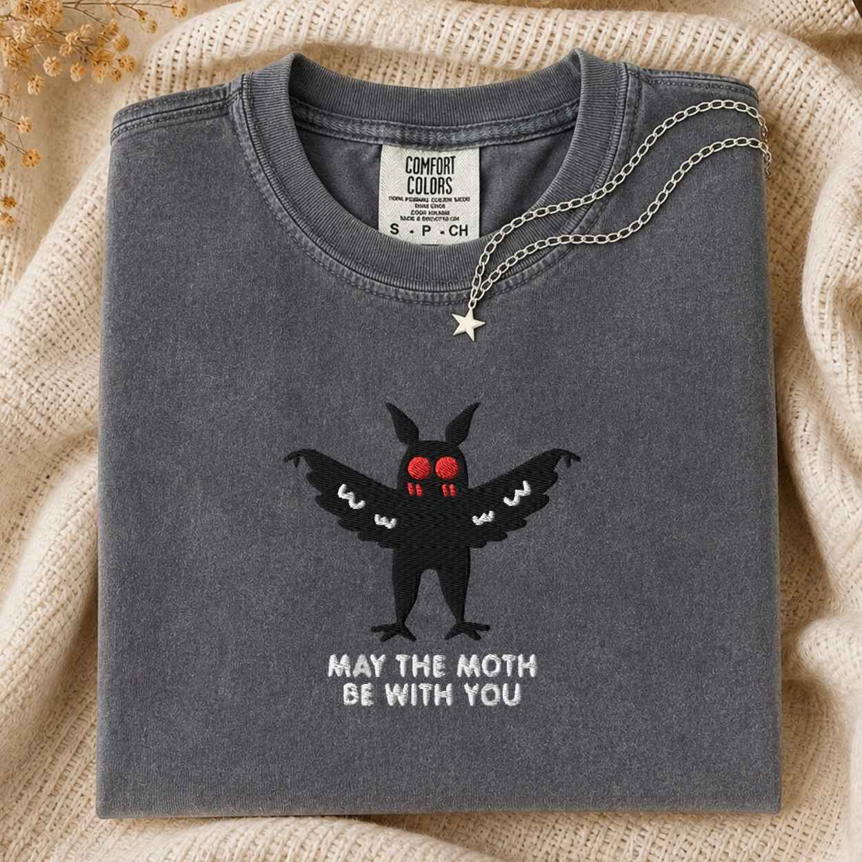 A folded charcoal gray Comfort Colors t-shirt featuring a black mothman graphic with red eyes and the text 'MAY THE MOTH BE WITH YOU' in white. A silver star pendant necklace is draped over the collar.