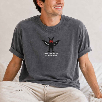 A man wearing a dark gray washed graphic t-shirt with a black moth design and the text 'MAY THE MOTH BE WITH YOU' on the chest, paired with beige pants, sitting on a bed.