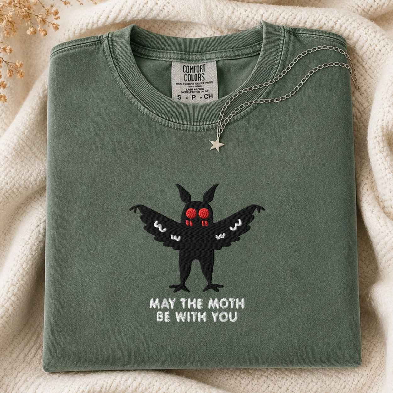 A folded sage green Comfort Colors t-shirt with a black moth graphic and the text 'MAY THE MOTH BE WITH YOU' on the front, styled on a cream knit blanket with a silver star necklace.
