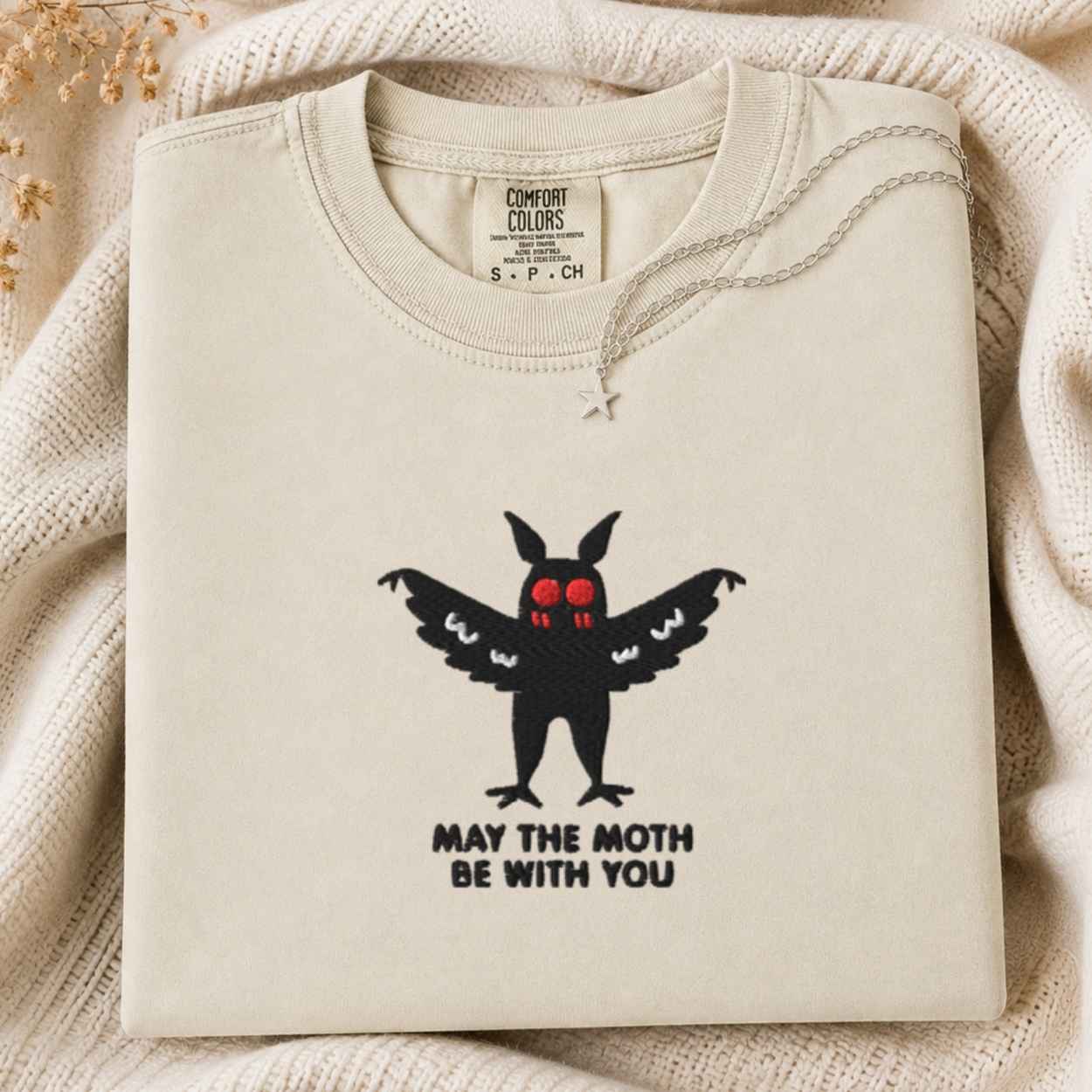 A folded cream sweatshirt with a black mothman graphic and the text 'MAY THE MOTH BE WITH YOU' on the front, displayed on a soft cream knit blanket. The inner neck label shows the brand 'Comfort Colors'.