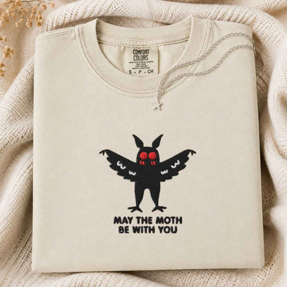 A folded cream sweatshirt with a black mothman graphic and the text 'MAY THE MOTH BE WITH YOU' on the front, displayed on a soft cream knit blanket. The inner neck label shows the brand 'Comfort Colors'.