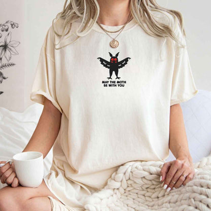A woman wearing a cream-colored oversized t-shirt with a black moth graphic and the text 'MAY THE MOTH BE WITH YOU' on the chest. She is sitting in a cozy room setting, holding a white mug and wrapped in a chunky knit blanket.