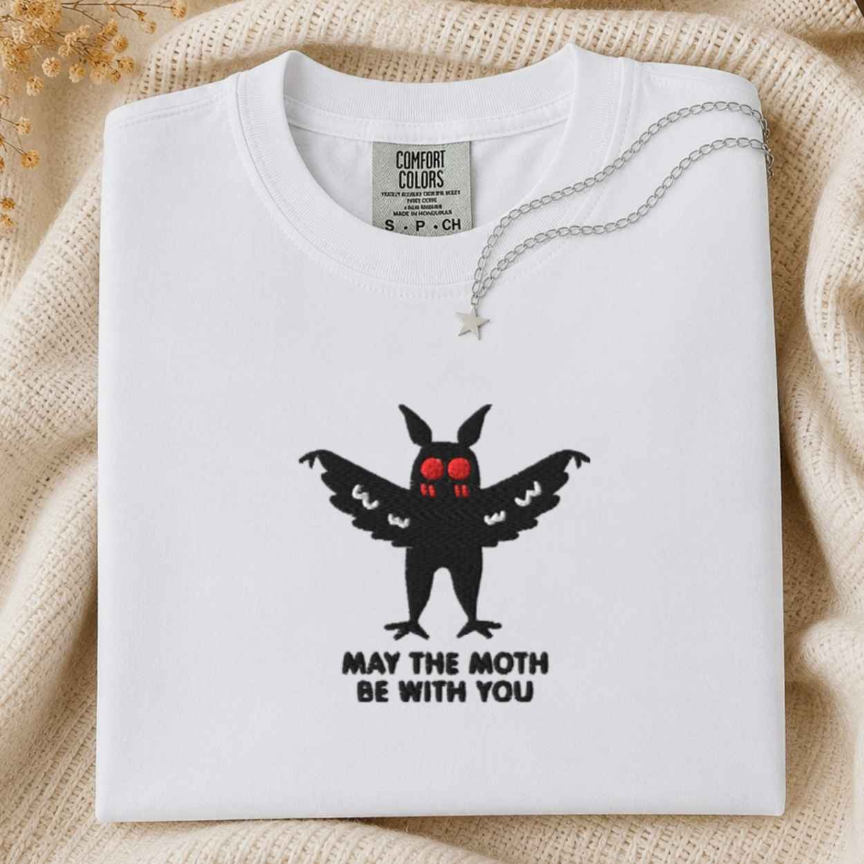 A folded white Comfort Colors t-shirt with a black moth graphic that has red eyes and the text 'MAY THE MOTH BE WITH YOU' underneath. A silver necklace with a star pendant is draped over the collar.