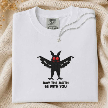 A folded white Comfort Colors t-shirt with a black moth graphic that has red eyes and the text 'MAY THE MOTH BE WITH YOU' underneath. A silver necklace with a star pendant is draped over the collar.