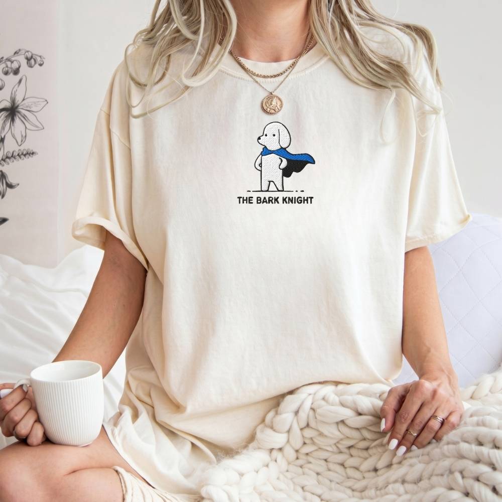 A woman wearing a cream-colored t-shirt with a graphic of a cartoon dog superhero and the text 'THE BARK KNIGHT'. She is sitting on a bed with a chunky knit blanket and holding a white mug.