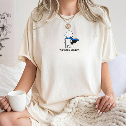 A woman wearing a cream-colored t-shirt with a graphic of a cartoon dog superhero and the text 'THE BARK KNIGHT'. She is sitting on a bed with a chunky knit blanket and holding a white mug.