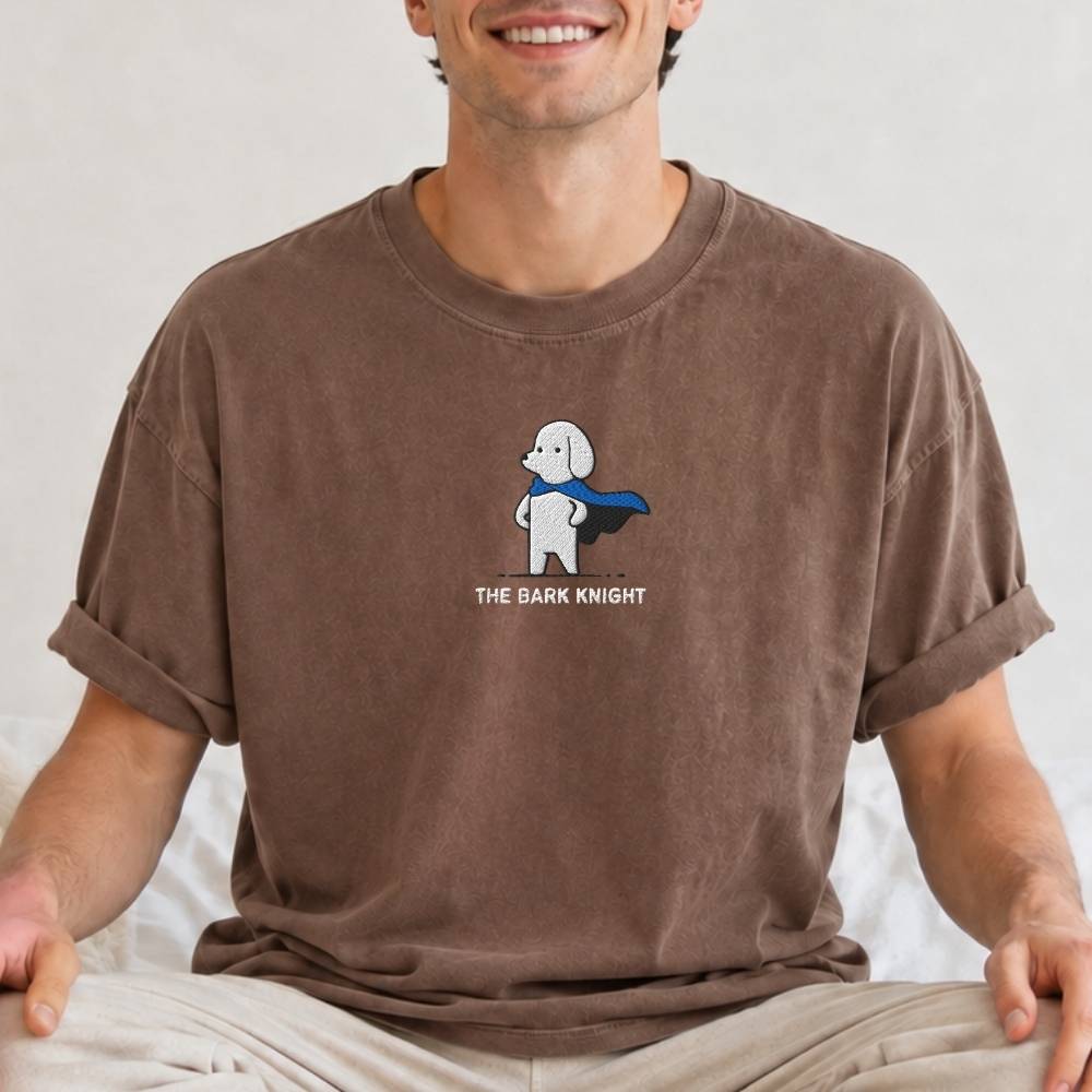 A man wearing a brown short-sleeve t-shirt with a cartoon graphic of a white dog in a blue cape and the text 'THE BARK KNIGHT' on the chest, sitting against a light background.