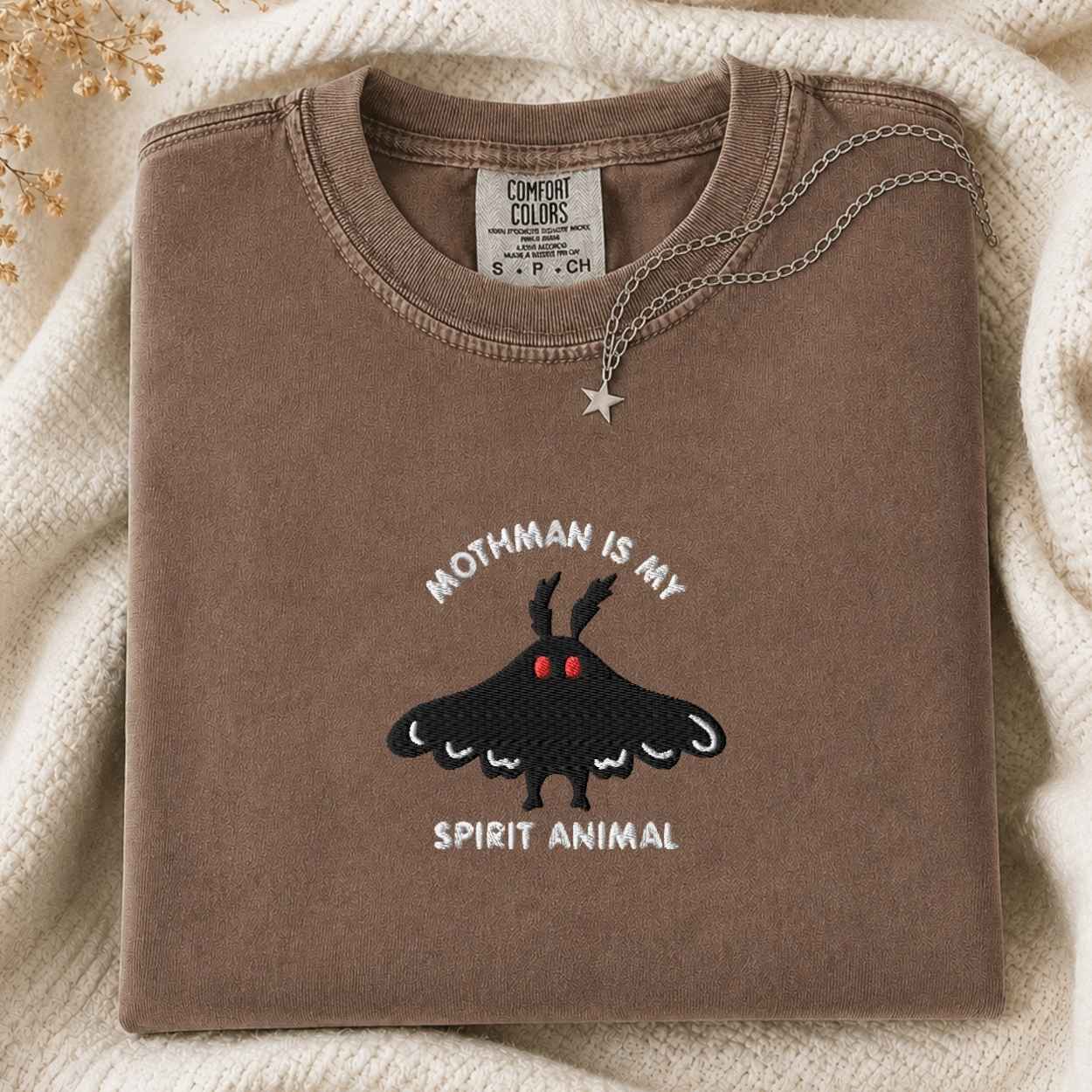 A folded brown t-shirt with a moth graphic and the text 'Mothman is my Spirit Animal' is styled on a cream knit blanket with a silver necklace.