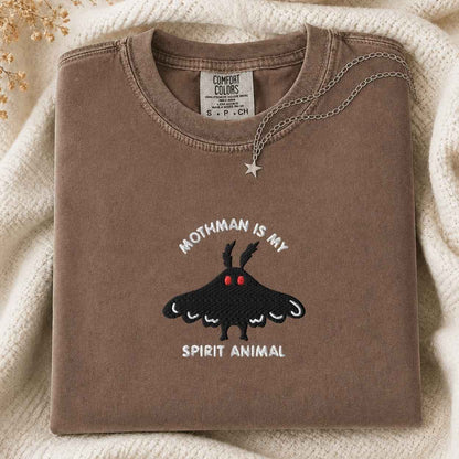 A folded brown t-shirt with a moth graphic and the text 'Mothman is my Spirit Animal' is styled on a cream knit blanket with a silver necklace.
