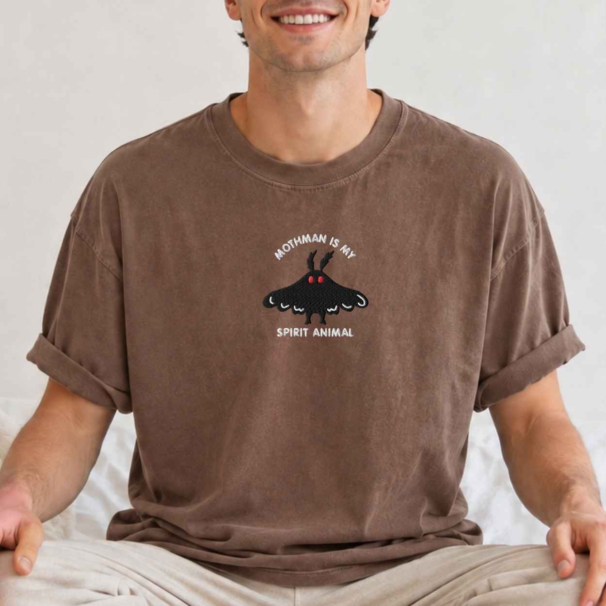 A man wearing a brown crewneck t-shirt with a graphic of a black moth and the text 'MOTHMAN IS MY SPIRIT ANIMAL' on the chest, sitting against a light background.