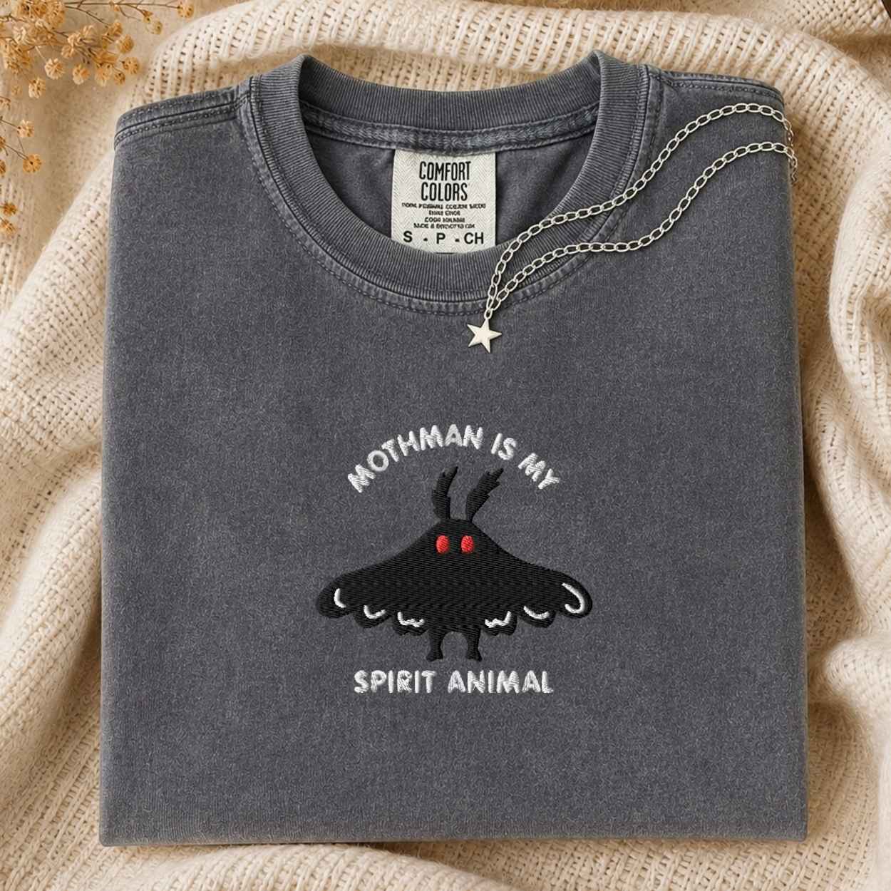 A folded dark gray t-shirt with a black moth graphic and the text 'MOTHMAN IS MY SPIRIT ANIMAL' on the front, styled on a beige knit blanket with a silver necklace draped over the collar.